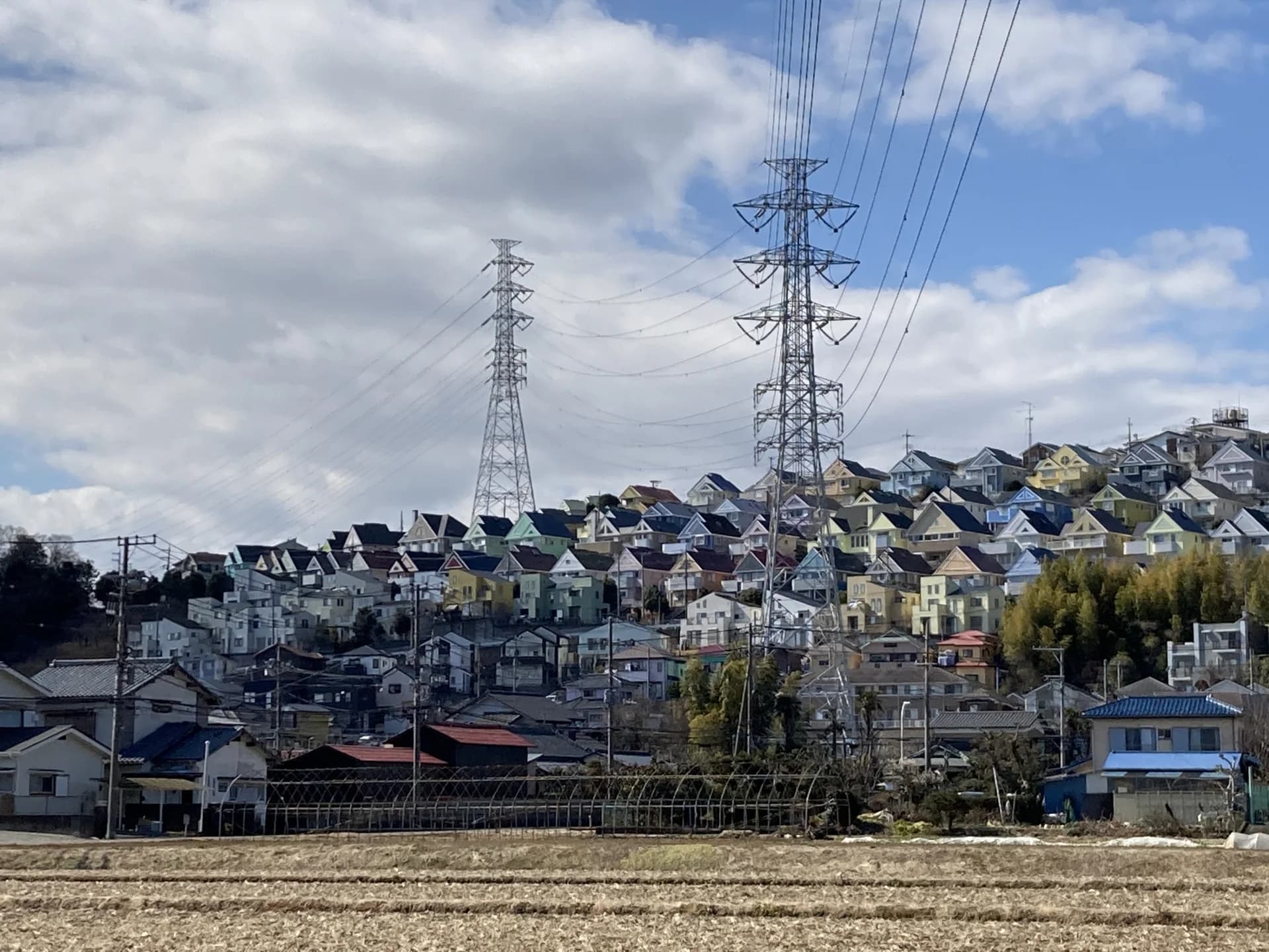畑の向こうの丘に戸建て住宅が建ち並び送電鉄塔が立つ平塚の街並み（平塚市の空き家買取のイメージ）