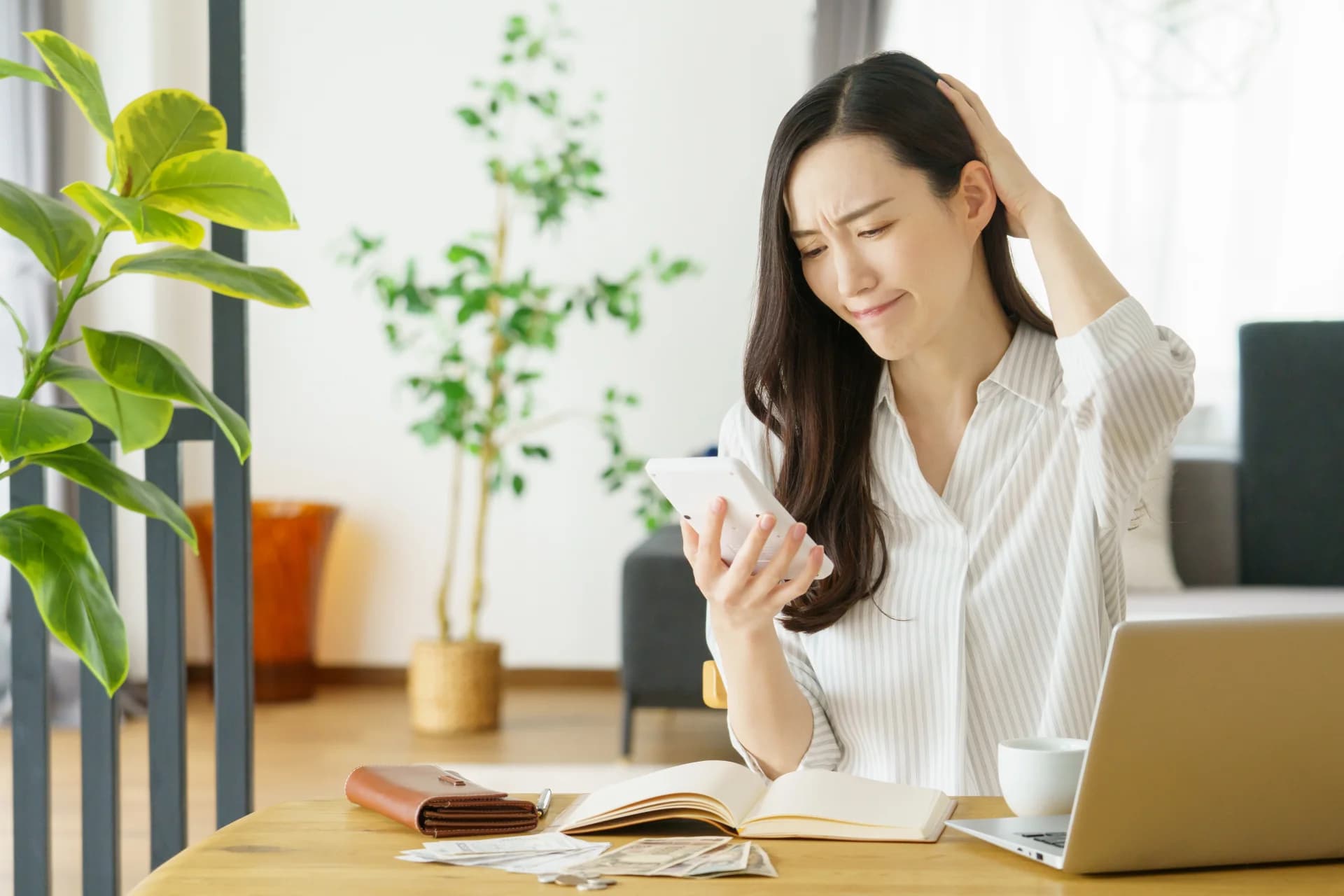 電卓を手にお札と家計簿を広げたテーブルの前で、困った顔で頭に手を当てる女性（家の解体費用を一度に払えず悩むイメージ）