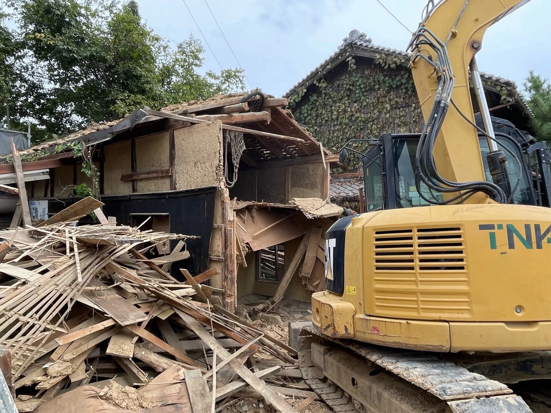 重機で取り壊し中の土壁の古い木造家屋と、奥に残るツタの絡まった家(広い敷地の家の解体のイメージ)