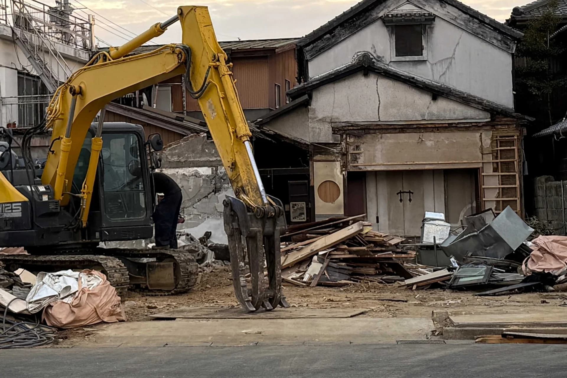 油圧ショベルで取り壊している途中の古いしっくい壁の家と、道路ぎわに積まれた廃材(土地を売る前の家の解体のイメージ)