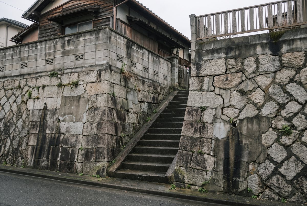 石積みの擁壁の上へ続く階段と、その脇に立つ古い住宅