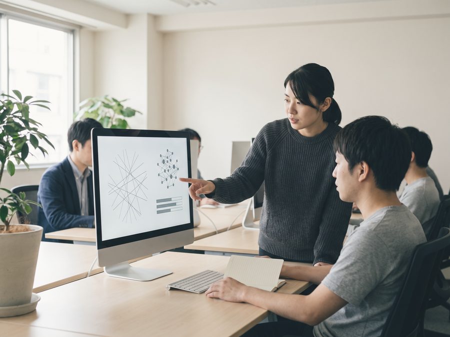 明るい執務フロアで、立った担当者が大きなモニターを指しながら、座った同僚と新しい事業の進め方を確かめている
