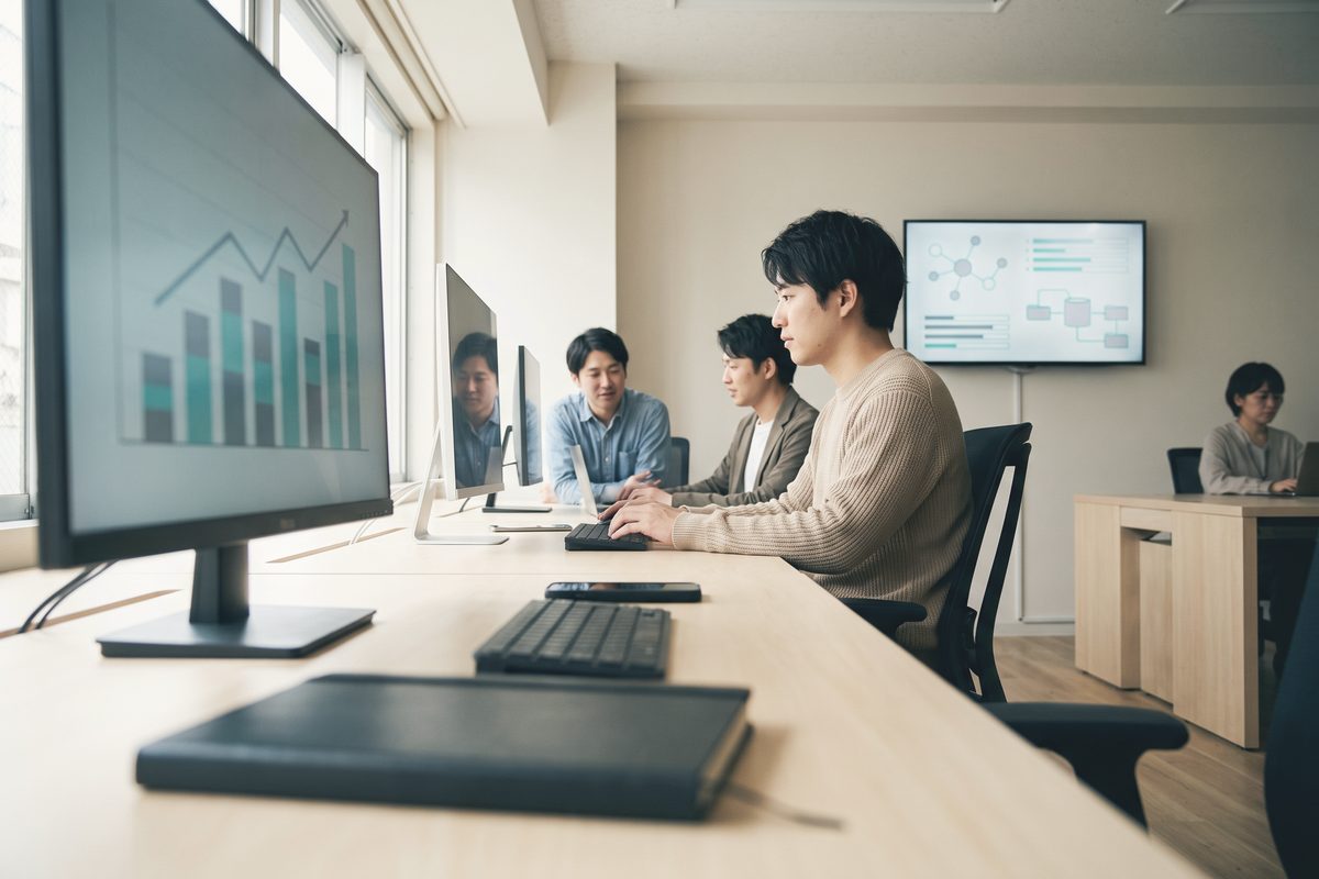 窓際の長机でキーボードを打つ人。手前のモニターに棒が並ぶ