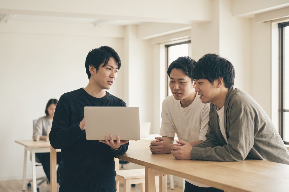 立ったままノートパソコンを抱え、画面を向けて説明している人と、机に手をついてのぞき込む2人