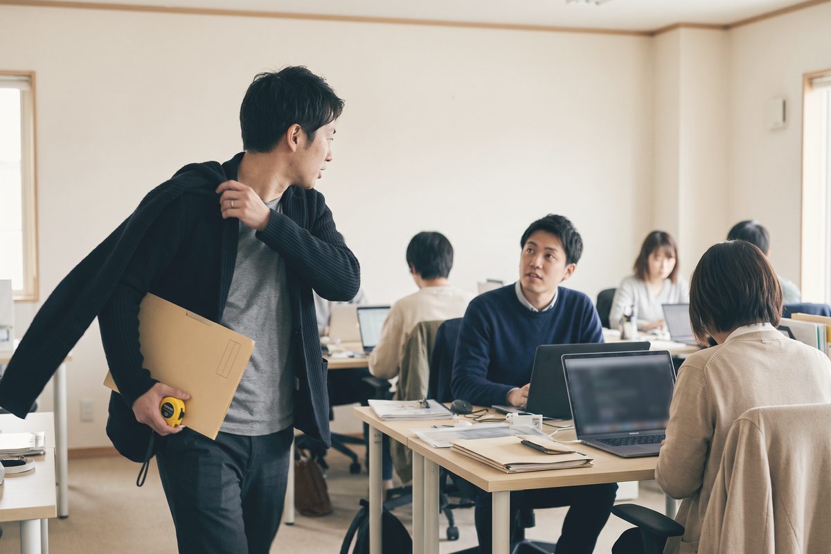 上着を肩に掛け、書類ばさみとメジャーを手に席を離れていく人。机の同僚が顔を上げて見送っている