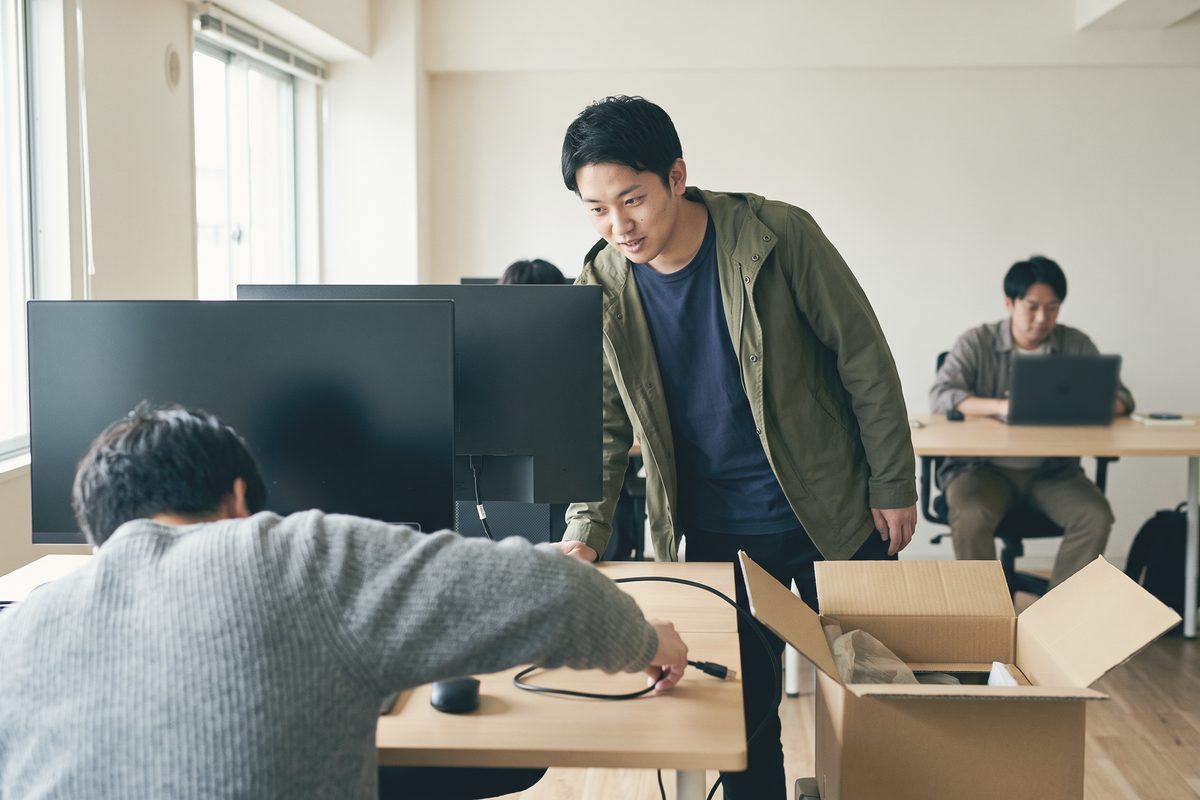 開けた段ボール箱の横で、自席に新しいモニターの配線をつないでいる人と、それを見に来た同僚
