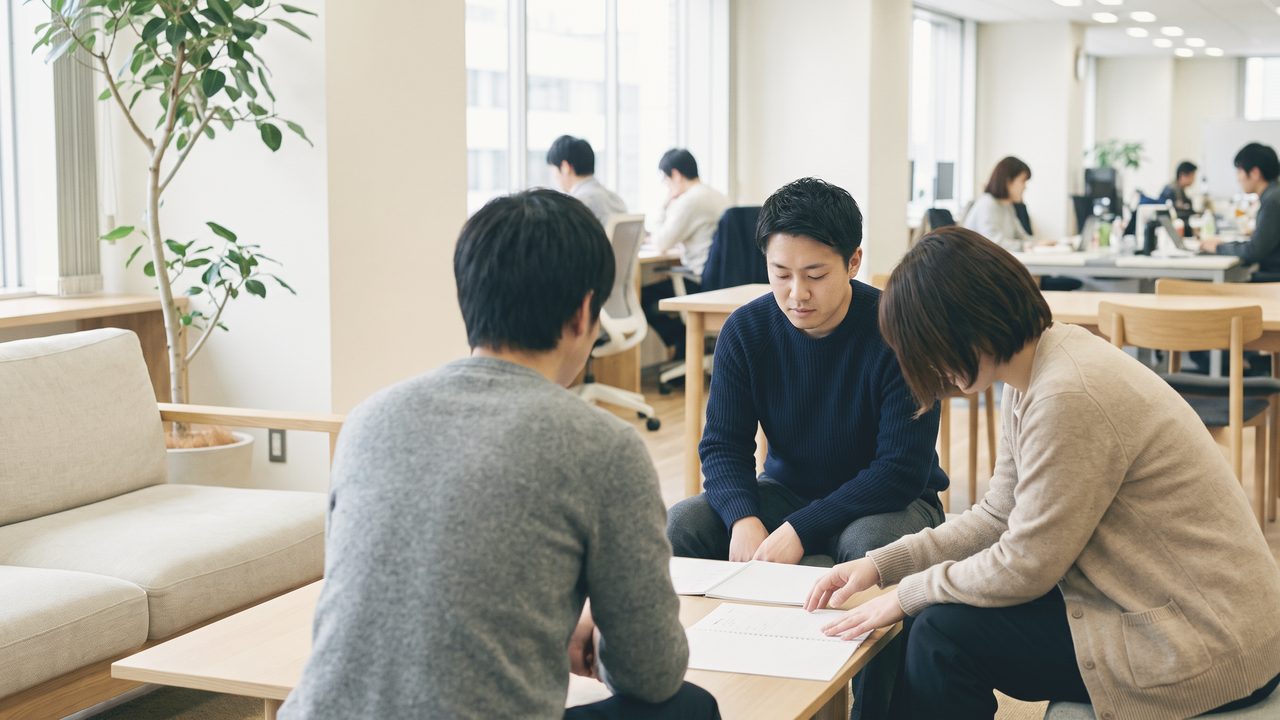 明るい執務フロアの一角で、3人が低い机を囲んで紙の資料を見ながら話している。奥では別の人たちが机に向かっている