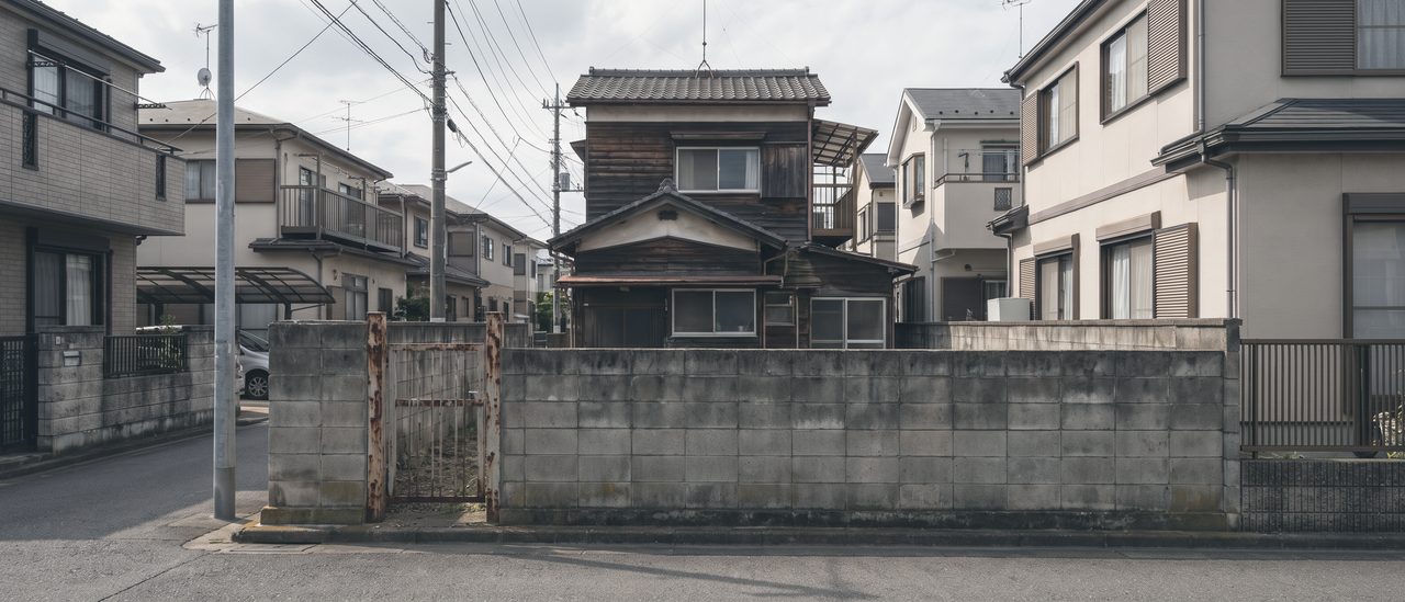 住宅街の道に面した低いブロック塀と門柱。その奥に、隣の家と近接して建つ古い2階建ての側面が見える