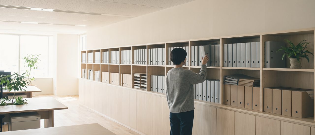 明るい執務フロアの壁際に並ぶ棚。同じ大きさのファイルと書類箱が隙間なく並び、手前で1人が背を向けて1冊を取り出している