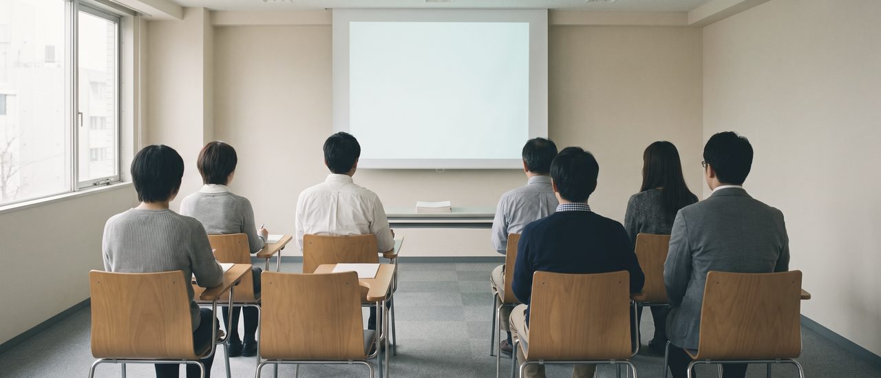 小さなセミナー室を最後列から見たところ。前を向いて座る数人の背中が並び、正面には何も映っていない明るい投影面がある