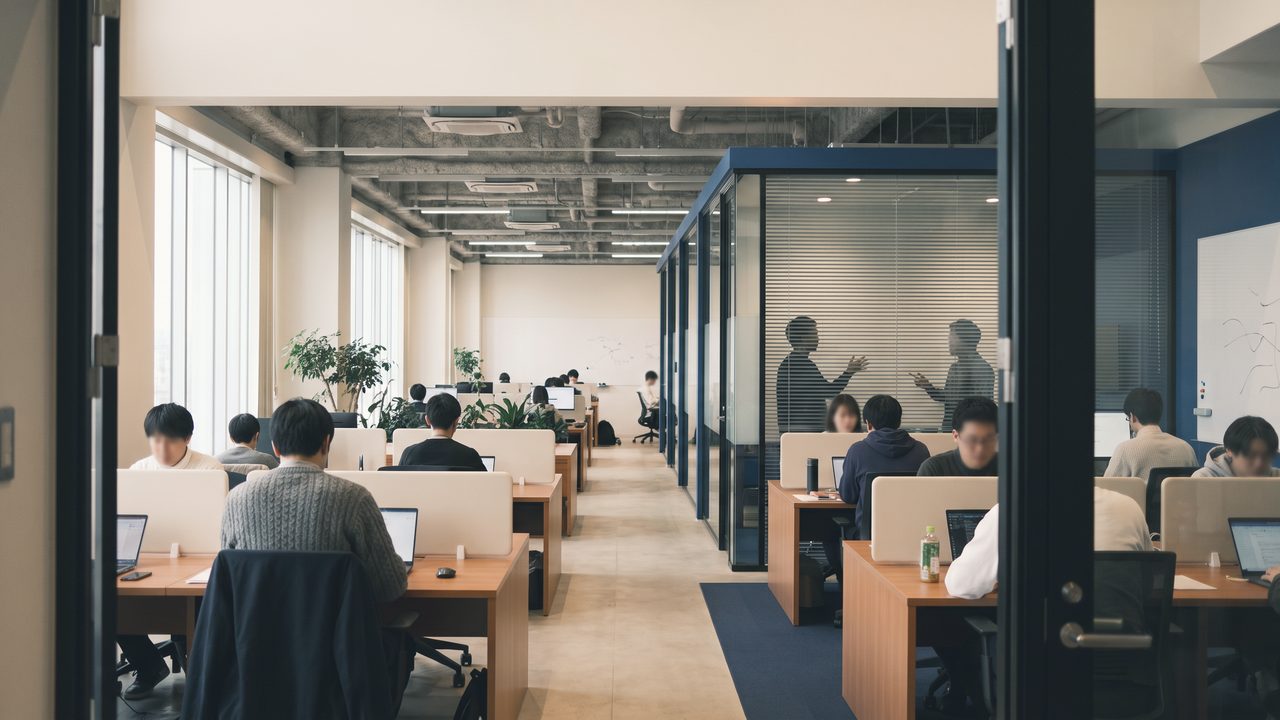 ガラスの会議室が並ぶ広いオフィスで、机に向かって作業している人たち