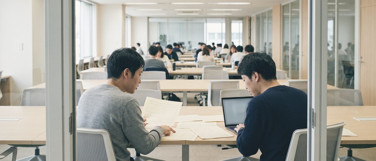 ガラスの仕切り越しに、机に並べた紙とノートパソコンをのぞき込み、片方が紙を指している2人