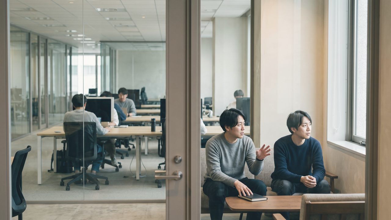 オフィスの一角で、机を挟んで向かい合って話している2人。ガラスの仕切り越しに、奥の執務フロアが見えている