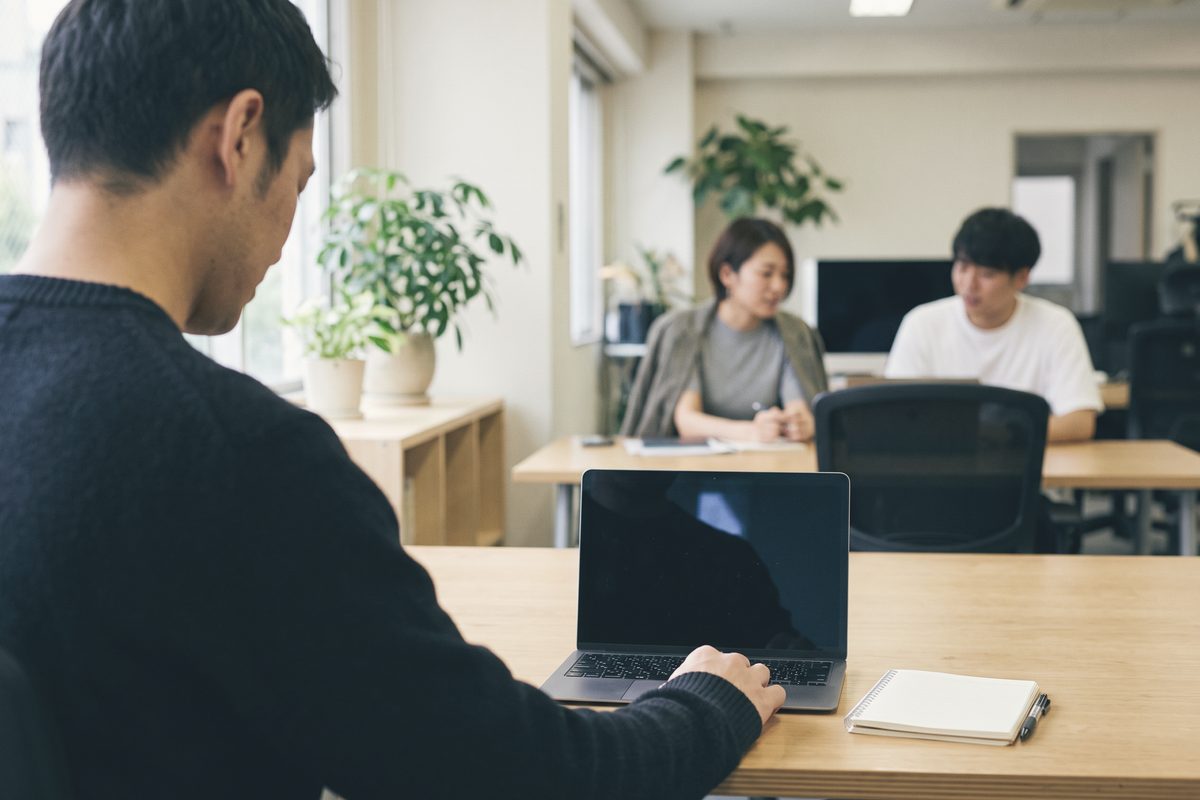 窓際の机でノートパソコンに向かう人の肩越しに見たところ。奥では2人が打ち合わせをしている