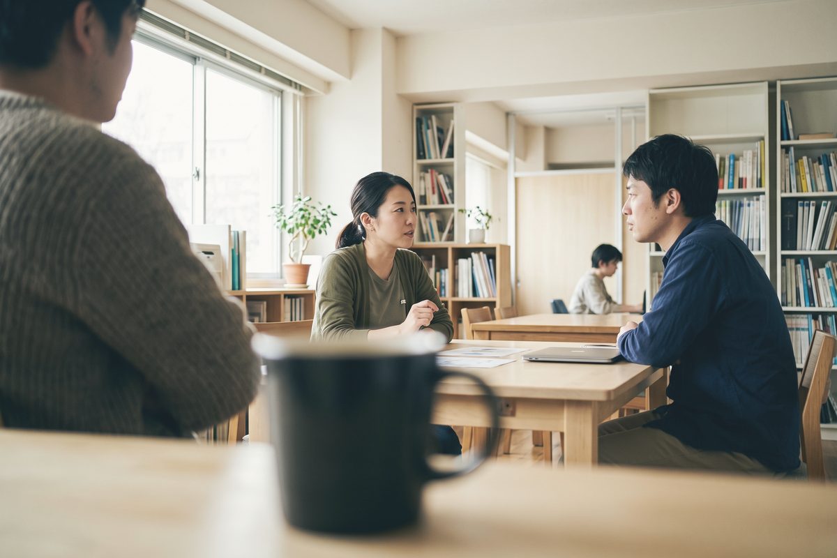 本棚のある小さな打ち合わせ室で、テーブルの向こうの2人が話している。手前にぼけた人の背中とマグカップ