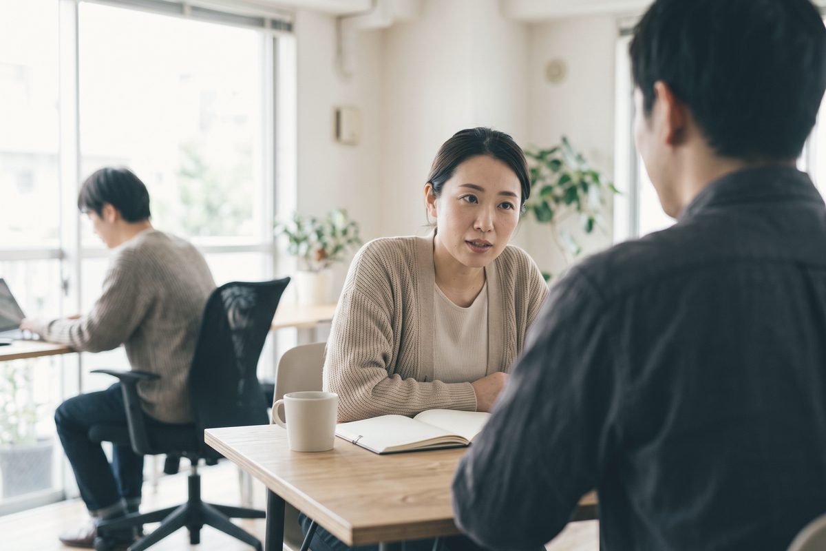 窓際の小さな木の机で、カーディガンの人が向かいの相手に身を乗り出して話している