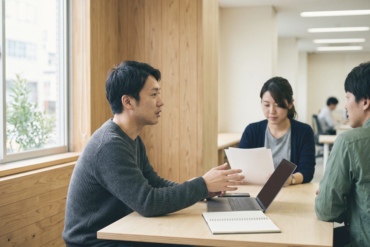木の壁の小さな部屋で、ノートパソコンを挟んで向かいの相手に説明している人
