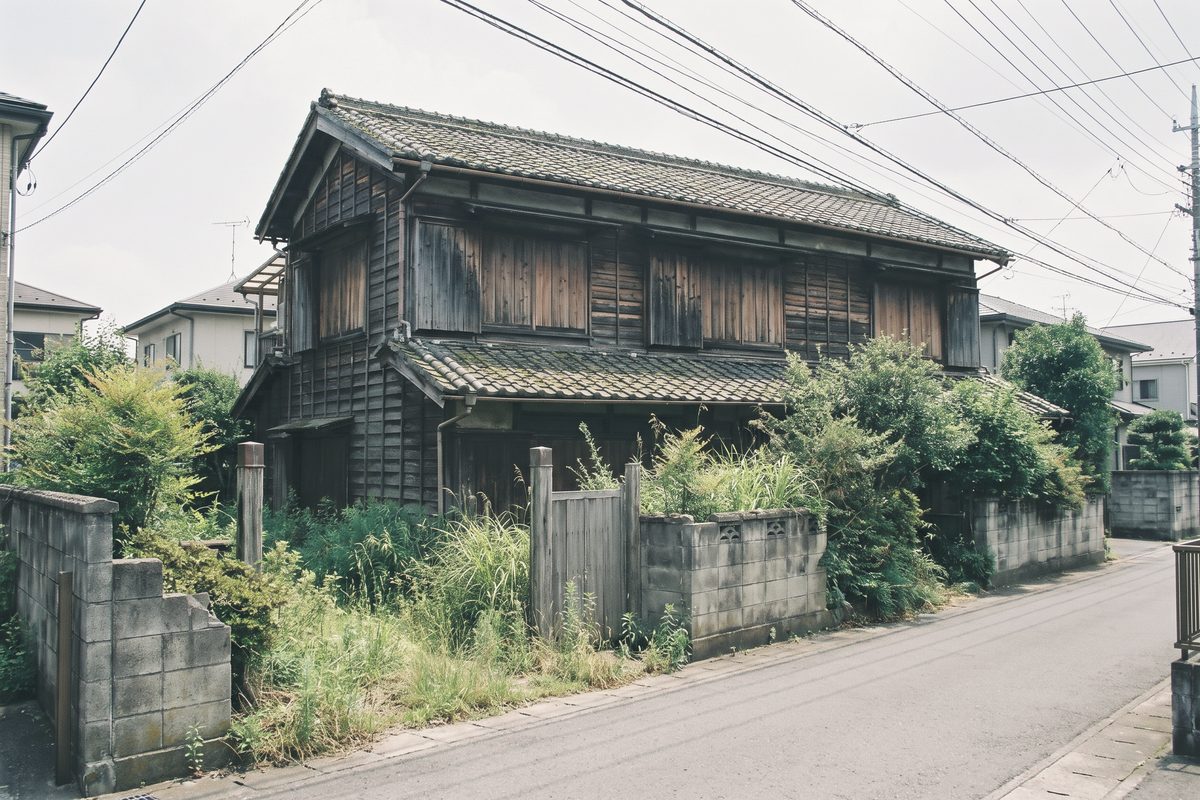 道の向かいから見た古い2階建て木造の空き家。雨戸は閉まり、ブロック塀と木の門の内側に草木が伸び放題になっている
