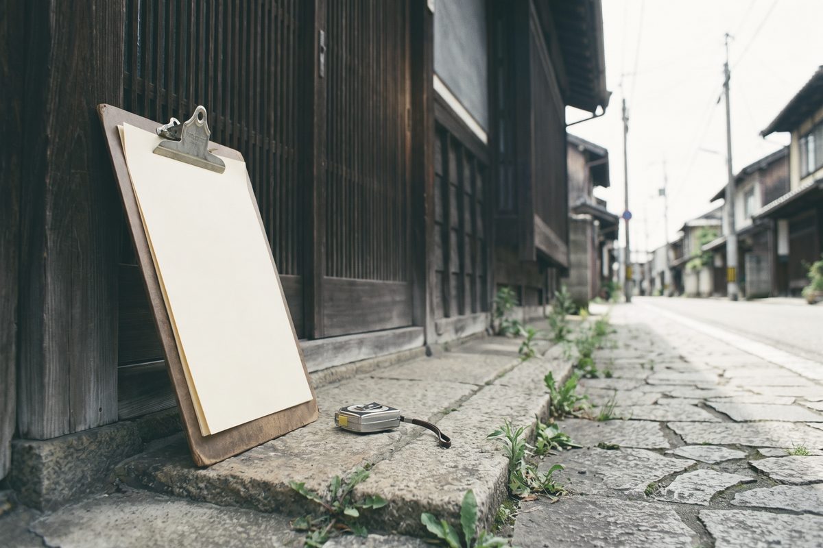 古い木造の家の玄関先に、白紙を挟んだクリップボードが立てかけてあり、横にメジャーが置いてある。草の生えた石畳の道が奥へ続いている