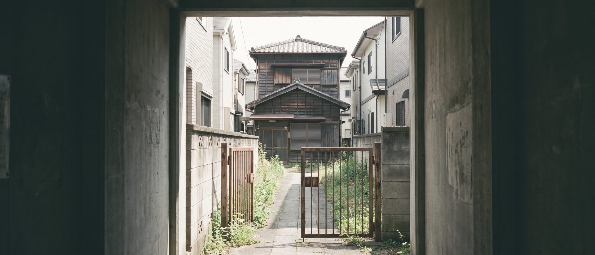 両側を家の壁に挟まれた細い通路の先に、古い木造2階建ての家が1軒だけ建っている。手前の錆びた門が半分開いている