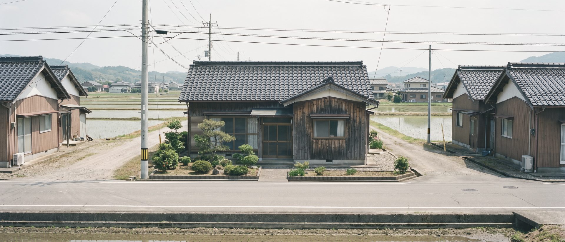 田んぼの広がる地方の町の道沿いに建つ、瓦屋根の小さな木造の平屋。両隣にも同じような古い家が並ぶ