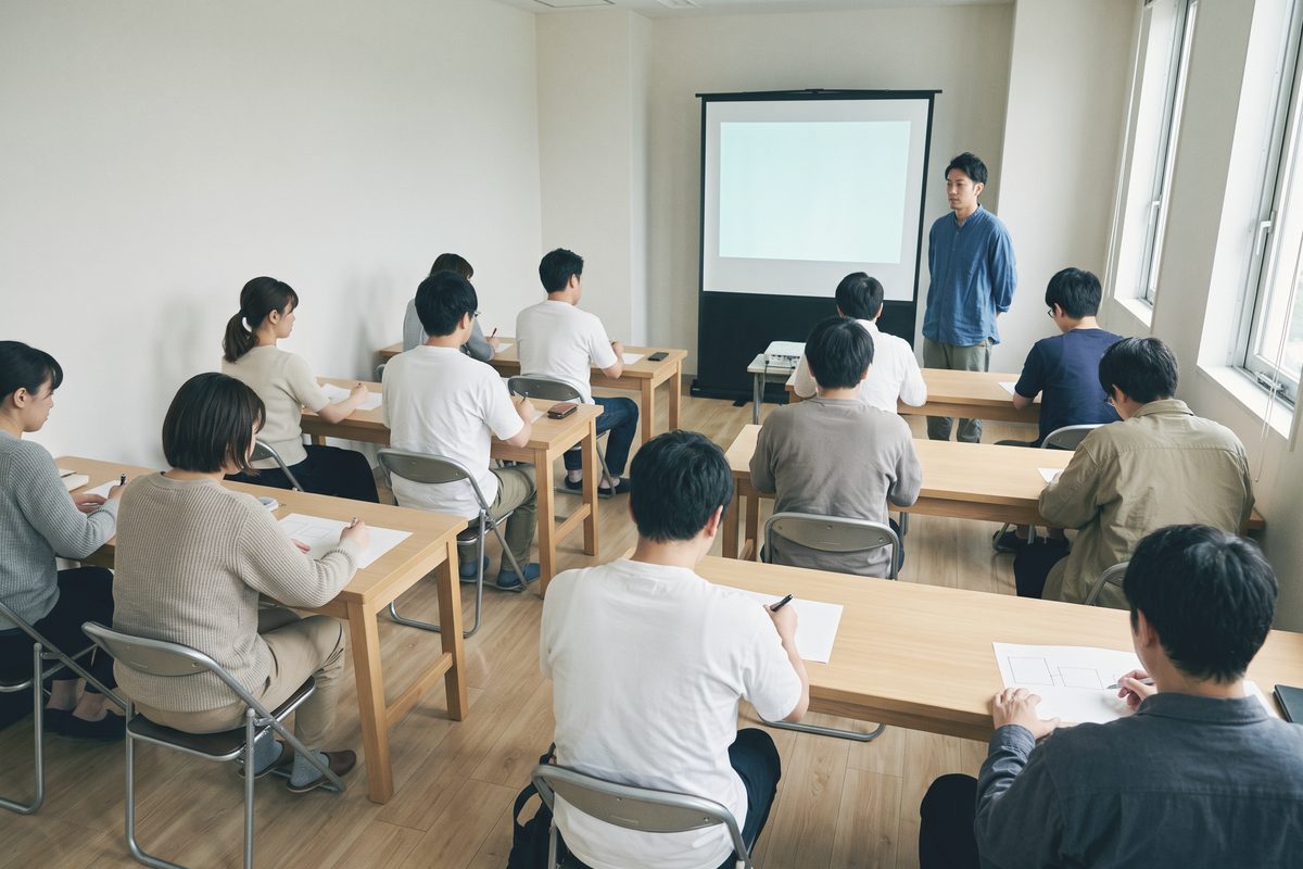 明るいセミナー会場を後ろから見たところ。10人ほどが机に向かい、前で1人が白い画面の横に立って話している
