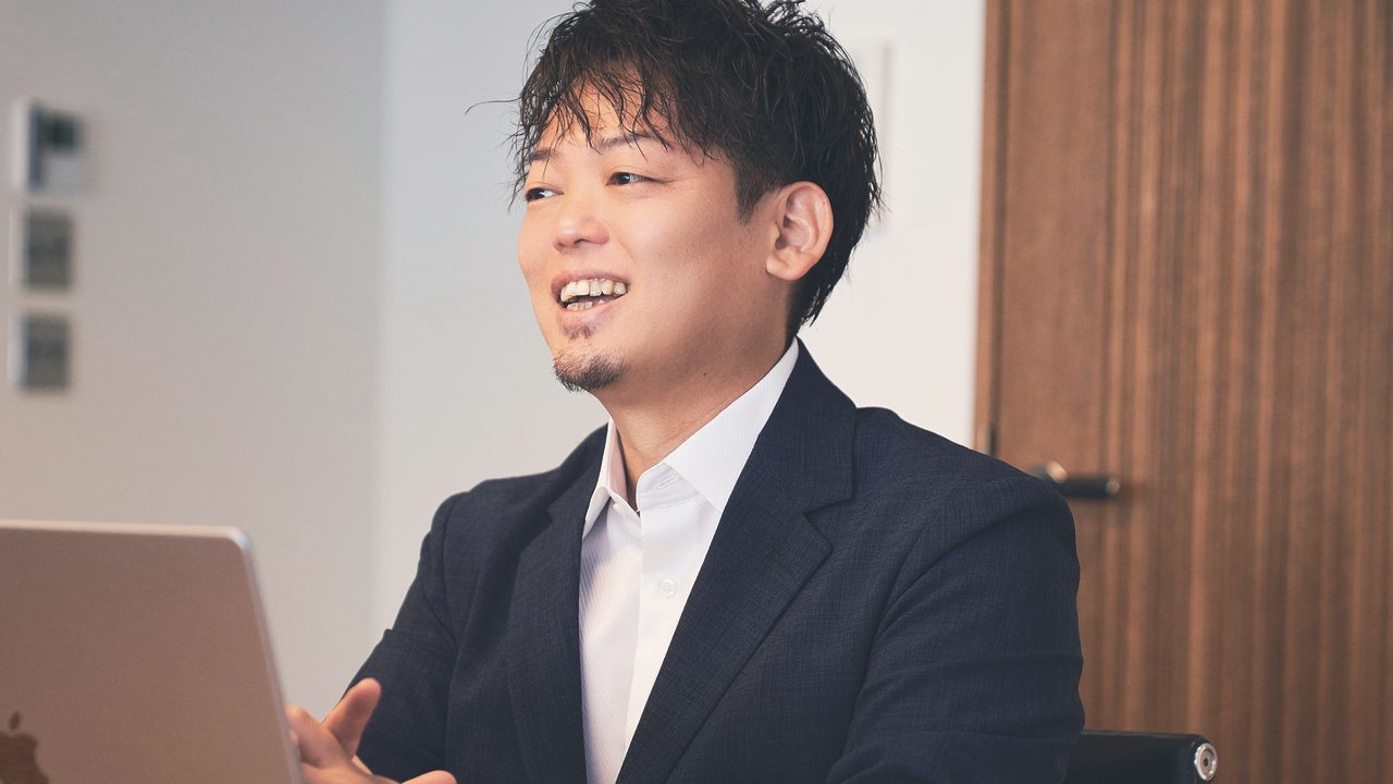 ノートパソコンを前に、机の向こう側の相手へ笑顔で話している藤山大二郎。背後は白い壁と木の扉
