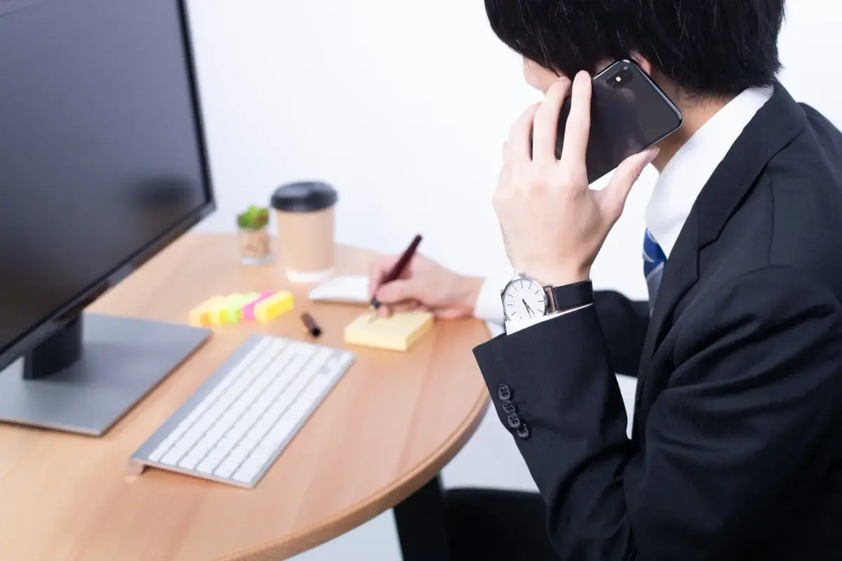 デスクトップPCの前で電話を取る日本人男性(不動産買取業者に関する記事のイメージ)