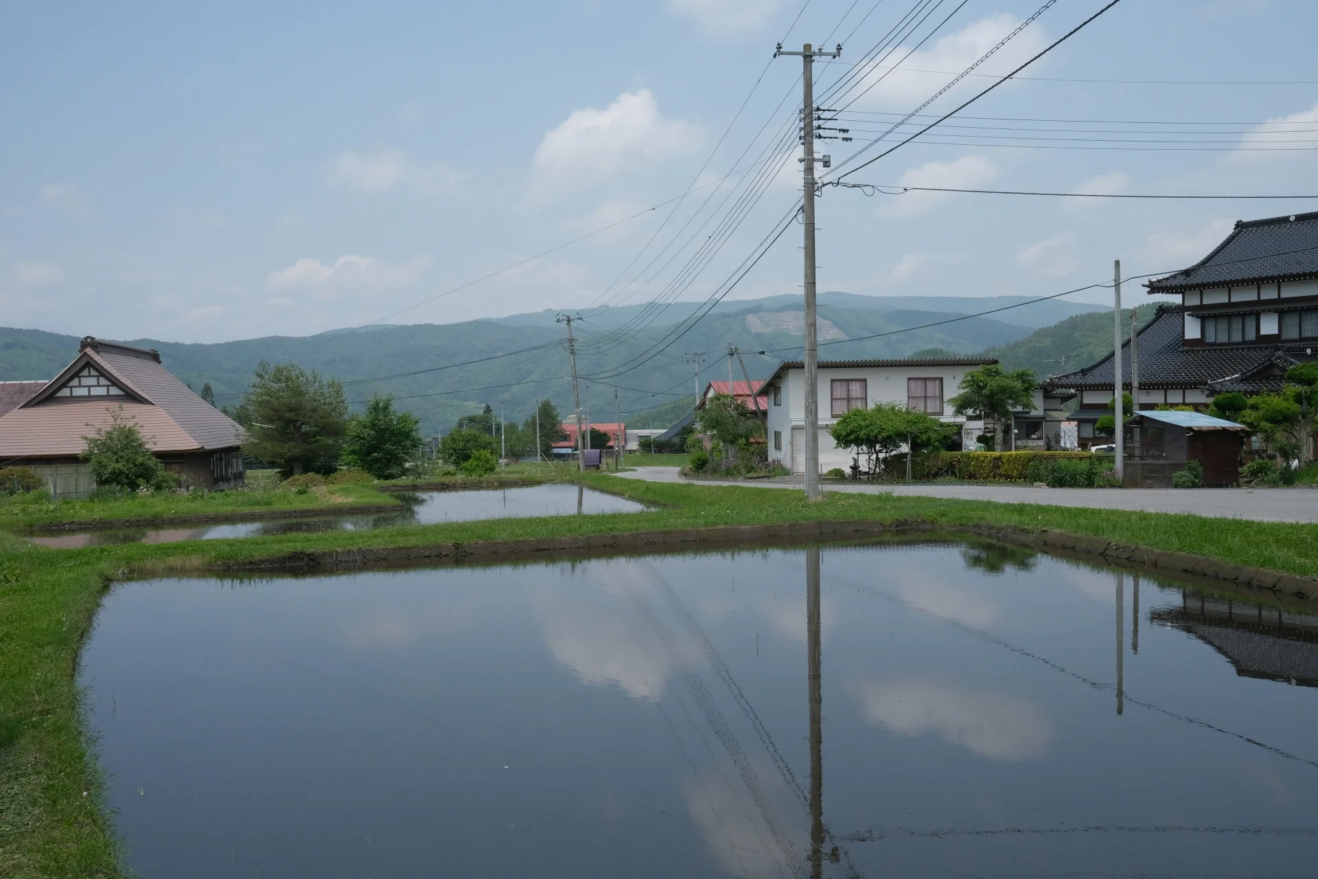 水を張った田んぼの奥に瓦屋根の家と山並みが見える初夏の田舎の町並み(名寄市の家の手放し方のイメージ)